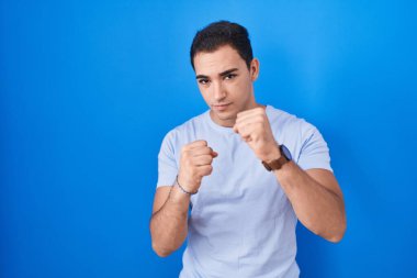 Young hispanic man standing over blue background ready to fight with fist defense gesture, angry and upset face, afraid of problem 