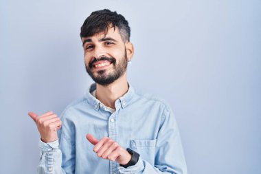 Mavi arka planda dikilen sakallı genç bir İspanyol. Arka tarafı işaret ediyor. El ve baş parmakları havada, kendinden emin. 