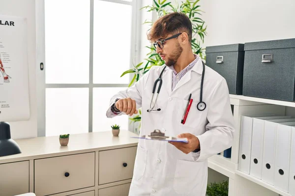 Young Arab Man Wearing Doctor Uniform Holding Clipboard Looking Watch — Stockfoto