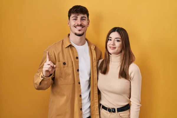 自信と幸せ笑みを浮かべている間 若いヒスパニック系のカップルは 黄色の背景が表示され 指の番号1で指摘します — ストック写真