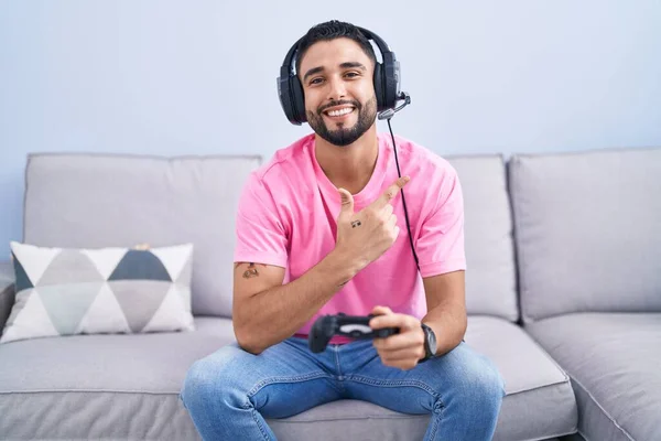 西班牙裔年轻人玩电子游戏 手里拿着控制器 坐在沙发上 面带微笑 手指手画脚地朝旁边看去 脸上带着快乐而自然的表情 — 图库照片