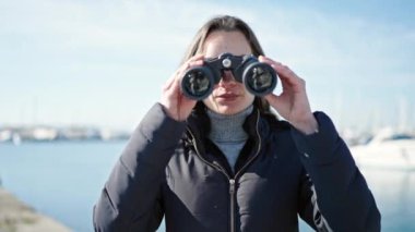 Genç beyaz kadın deniz kenarında dürbün kullanıyor.