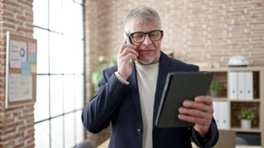 Middle age grey-haired man business worker talking on smartphone using touchpad at office