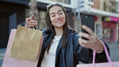 Genç ve güzel İspanyol kadın, elinde alışveriş torbalarıyla akıllı telefonuyla selfie çekiyor.