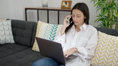 Genç, güzel, İspanyol bir kadın akıllı telefonuyla evde dizüstü bilgisayar kullanıyor.