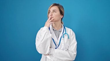 Genç sarışın kadın doktor şüpheli bir ifadeyle dikiliyor ve mavi arka planı düşünüyor.