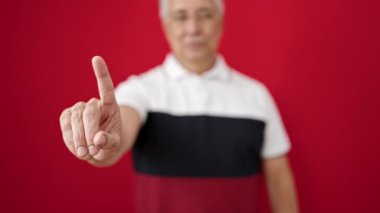 Gri saçlı orta yaşlı bir adam, izole edilmiş kırmızı arka planda parmağıyla hayır diyor.