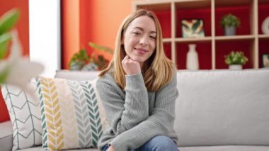 Sarışın genç bir kadın evdeki koltukta kendinden emin bir şekilde oturarak gülümsüyor.