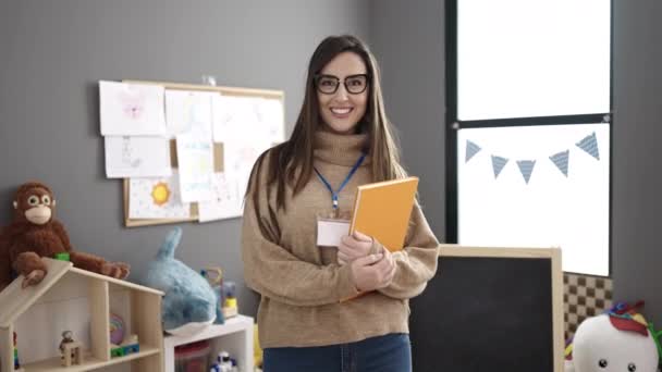 Mooie Spaanse Vrouw Kleuterleidster Glimlachend Zelfverzekerd Holding Boek Kleuterschool — Stockvideo