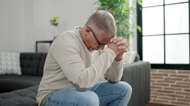 Orta yaşlı, kır saçlı, stresli bir adam evdeki koltukta oturuyor.