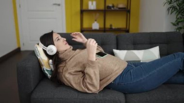 Evdeki kanepede uzanmış akıllı telefonuyla müzik dinleyen güzel İspanyol kadın.