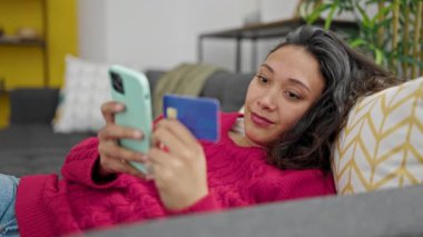 Genç, güzel, İspanyol bir kadın akıllı telefon ve kredi kartı alıp evdeki kanepede yatıyor.