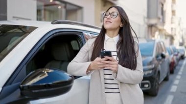 Genç, güzel, İspanyol bir kadın akıllı telefon kullanıyor. Sokakta arabaya yaslanıyor.