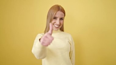 Sarışın genç bir kadın, izole edilmiş sarı arka planda baş parmağıyla gülümsüyor.