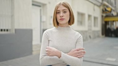 Ciddi bir ifade ve kollarıyla caddede duran genç sarışın kadın.