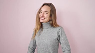 Genç, beyaz bir kadın, pembe arka planda kendine güvenen bir şekilde kameraya bakıyor.