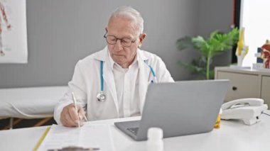 Kıdemli gri saçlı doktor klinikte boyun ağrısı şikayetiyle sağlık raporu yazıyor.