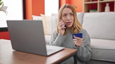 Akıllı telefondan konuşan genç sarışın kadın elinde kredi kartıyla evdeki kanepede oturuyor.
