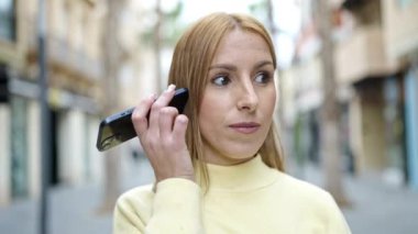 Young blonde woman smiling confident listening audio message by the smartphone at street