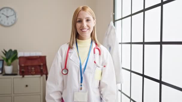 Νεαρή Ξανθιά Γυναίκα Γιατρός Χαμογελά Αυτοπεποίθηση Κρατώντας Πρόχειρο Στην Κλινική — Αρχείο Βίντεο