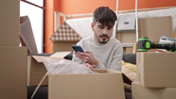 Joven Hombre Hispano Desempacando Caja Cartón Usando Smartphone Nuevo Hogar — Vídeo de stock