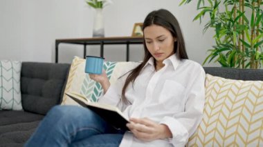 Young beautiful hispanic woman reading book and drinking coffee sitting on sofa at home