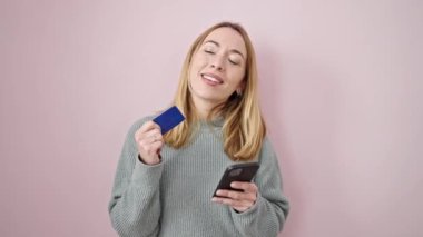 Akıllı telefon ve kredi kartıyla izole edilmiş pembe arka planda alışveriş yapan genç sarışın kadın.
