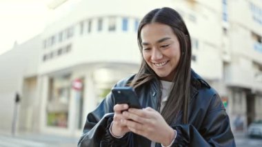 Genç, güzel, İspanyol bir kadın sokakta akıllı telefon kullanarak gülümsüyor.