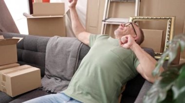Orta yaşlı, kır saçlı bir adam yeni evinde koltukta oturup kollarını esnetiyor.