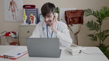 Genç İspanyol doktor bilgisayarını kullanarak klinikte telefonla konuşuyor.