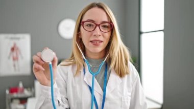 Klinikte steteskop tutan genç sarışın doktor gülümsüyor.