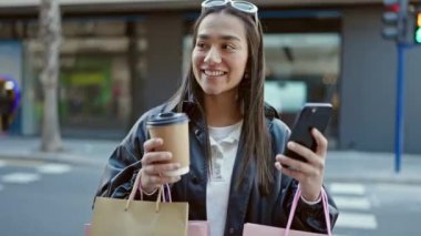 Genç ve güzel İspanyol kadın akıllı telefon kullanıyor. Elinde alışveriş poşetleri ve sokakta kahve tutuyor.
