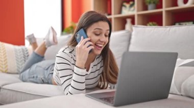 Young beautiful hispanic woman using laptop talking on smartphone at home