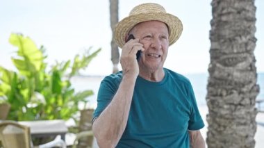 Kıdemli kır saçlı turist. Yazlık şapka takıyor. Deniz kenarında akıllı telefondan konuşuyor.
