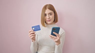 Akıllı telefon ve kredi kartıyla izole edilmiş pembe arka planda alışveriş yapan genç sarışın kadın.