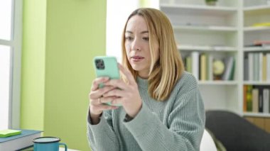 Kütüphanede akıllı telefon kullanan genç sarışın kız öğrenci.