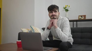 Genç İspanyol adam evdeki koltukta dizüstü bilgisayar kullanıyor.