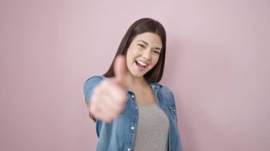 Genç, güzel, İspanyol bir kadın pembe arka planda baş parmağıyla gülümsüyor.