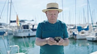 Limanda akıllı telefon kullanarak yaz şapkası takan yaşlı, kır saçlı bir turist.