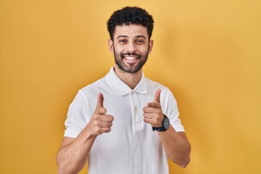 Sarı arka planda duran Arap adam mutlu ve komik yüzlü parmaklarını kameraya doğrultuyor. iyi enerji ve titreşimler. 