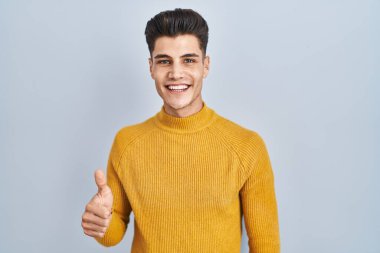 Mavi arka planda dikilen İspanyol genç adam elleriyle mutlu bir jest yapıyor. Kameraya bakıp başarıyı gösteren ifade. 