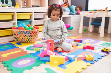 Adorable hispanic girl playing with construction blocks sitting on floor at kindergarten