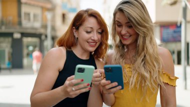 Two women standing together using smartphones at street