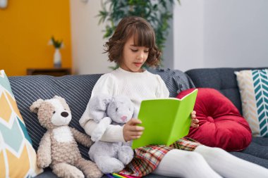Adorable hispanic girl reading book sitting on sofa at home