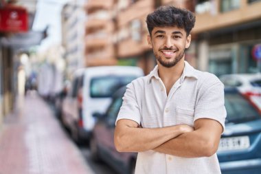 Genç Arap adam gülümsüyor. Kollarını kavuşturup caddede duruyor.