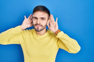 Hispanic man standing over blue background trying to hear both hands on ear gesture, curious for gossip. hearing problem, deaf 