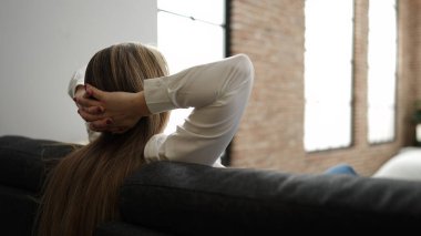 Sarışın genç kadın ellerini başının üstüne koyup evdeki kanepede oturuyor.