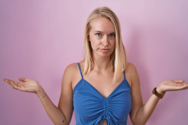 Young caucasian woman standing over pink background clueless and confused with open arms, no idea concept. 