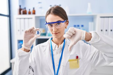 Genç, beyaz bir kadın laboratuvarda çalışıyor. Elinde kızgın yüzle Bitcoin tutuyor. Negatif işaretler baş parmakları aşağıda, reddedilme konsepti ise reddedilmiş. 