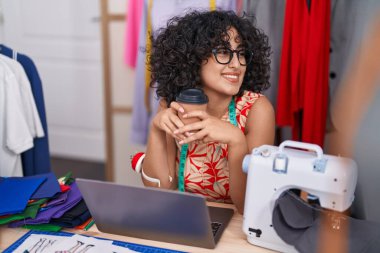 Young middle eastern woman tailor using laptop drinking coffee at tailor shop